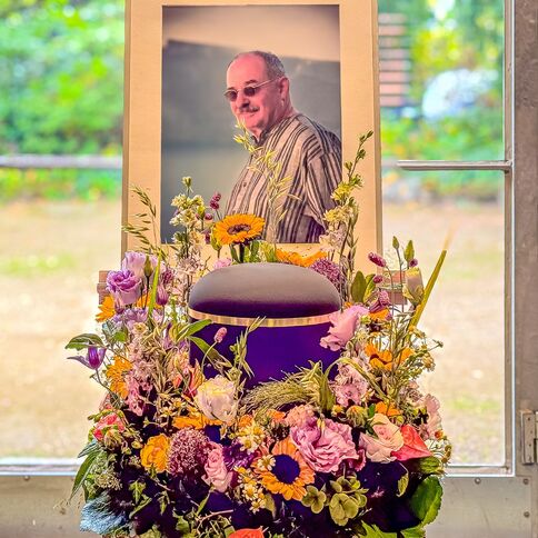 Ein Porträt von Stevens steht hinter einer Urne, umgeben von einem bunten Blumenarrangement in einem hellen Raum.