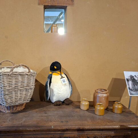 Ein Plüschpinguin steht auf einem Holztisch in Tuppenhof, umgeben von Körben und dekorativen Töpfen.
