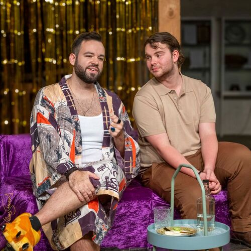 Zwei Männer sitzen in einem Theaterverein auf einem lila Sofa und führen ein lebhaftes Gespräch.