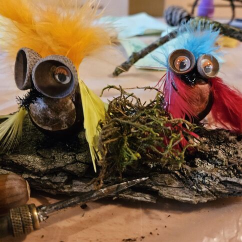 Zwei aus Naturmaterialien gebastelte Figuren mit bunten Federn sitzen auf einem Stück Rinde beim Herbstbasteln.