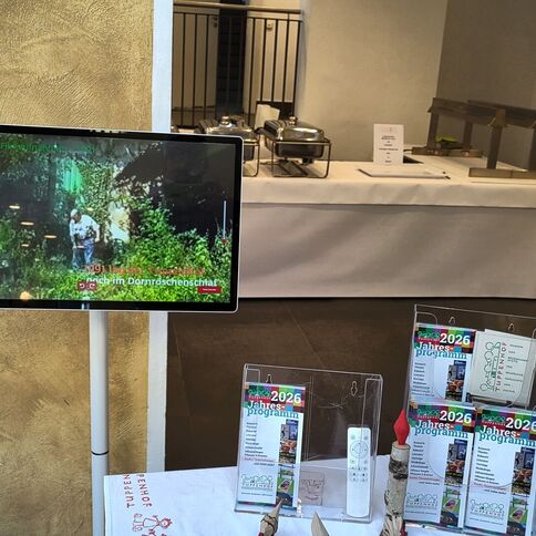 Ein Monitor zeigt Aktivitäten im Tuppenhof Kaarst, während im Hintergrund ein Buffet vorbereitet wird.