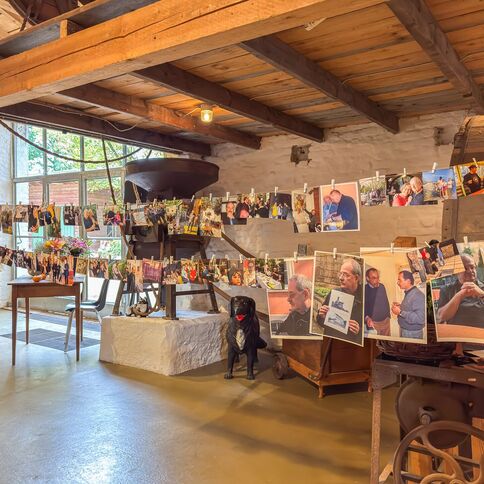 In einer rustikalen Scheune hängen zahlreiche Fotos an Leinen, während ein schwarzer Hund darunter sitzt.