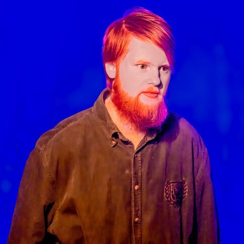 Ein Mann mit rotem Bart steht vor einem intensiven blauen Hintergrund, möglicherweise bei einer Veranstaltung im Tuppenhof.