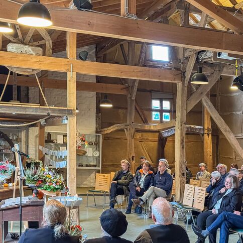Eine Frau hält in einem rustikalen Saal mit Holzbalken eine Rede vor einem aufmerksam lauschenden Publikum.