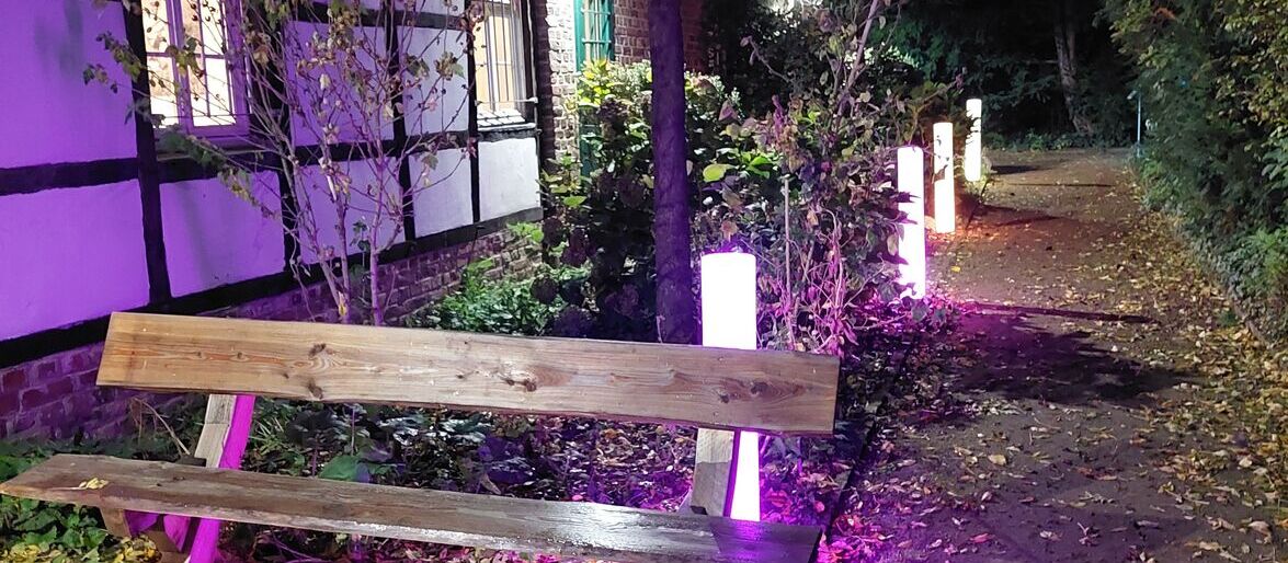 Eine Bank steht im Abendlicht neben einem Fachwerkhaus, umgeben von bunten Lichtern auf einem Laubweg.