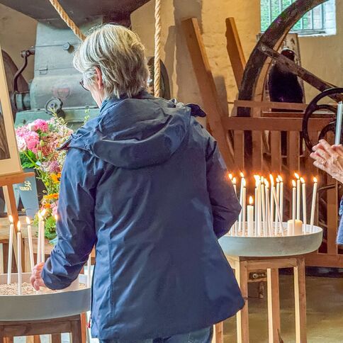 Zwei Personen stehen in einer Gedenkfeier vor einem Foto, umgeben von brennenden Kerzen und Blumen.
