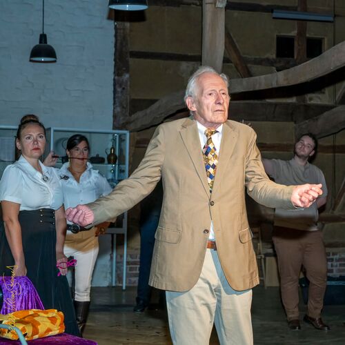 Ein älterer Mann in einem Anzug steht auf einer Bühne, während andere Personen im Hintergrund aufmerksam zuschauen.