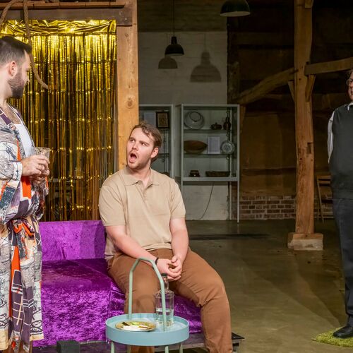 Drei Männer führen in einem Theaterstück einen lebhaften Dialog vor glitzerndem Hintergrund.