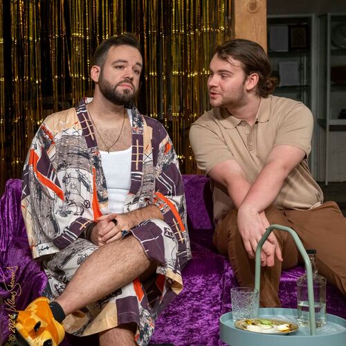 Zwei Männer sitzen in einem gemütlichen Raum auf einem lila Sofa und unterhalten sich angeregt.
