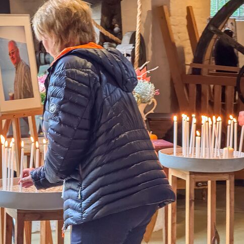 Zwei Personen zünden Kerzen an, während ein Porträt von Stevens auf einer Staffelei im Hintergrund steht.