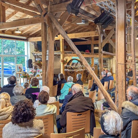 Eine Gruppe von Menschen sitzt in einem rustikalen Saal und hört einer Rednerin an einem Pult zu.