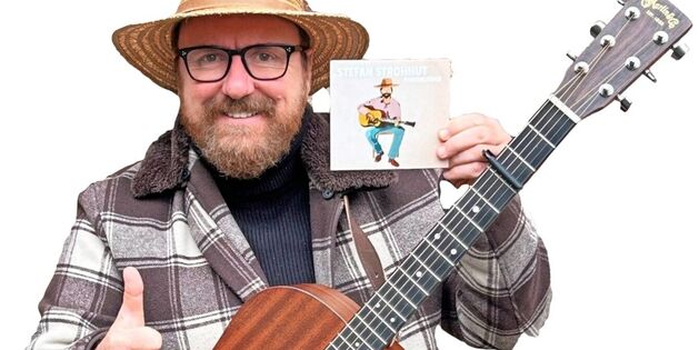 Ein Mann mit Strohhut hält eine Akustikgitarre und eine CD, während er lächelnd den Daumen hebt.
