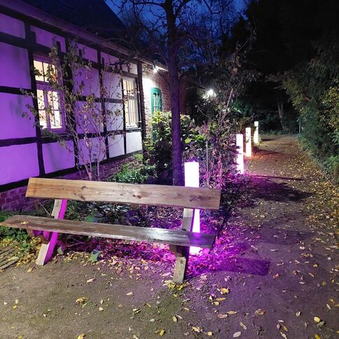 Eine Bank steht im Abendlicht neben einem Fachwerkhaus, umgeben von bunten Lichtern auf einem Laubweg.