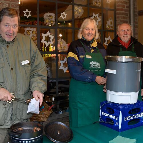 Drei Personen stehen lächelnd an einem Weihnachtsstand und servieren heiße Getränke in festlicher Atmosphäre.
