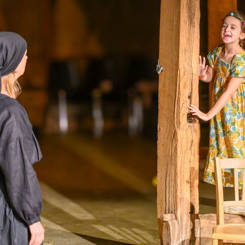 Ein Kind im bunten Kleid spielt fröhlich auf einem Holzbalken, während eine Frau in traditioneller Kleidung zuschaut.