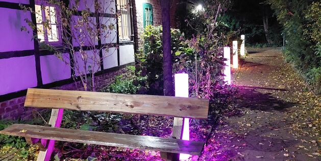 Eine Bank steht im Abendlicht neben einem Fachwerkhaus, umgeben von bunten Lichtern auf einem Laubweg.