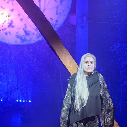Eine Person mit langer weißer Perücke steht auf einer Bühne in Kaarst, beleuchtet von mystischem blauem Licht im Tuppenhof.