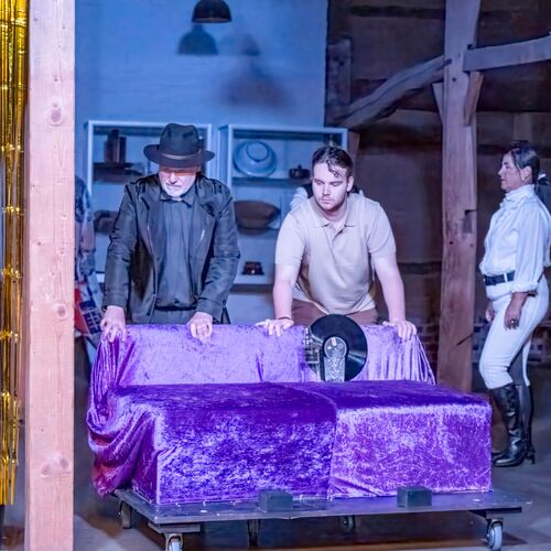 Zwei Männer in einem Theaterstück schieben einen mit lila Stoff bedeckten Wagen, während eine Frau in Reitkleidung zusieht.