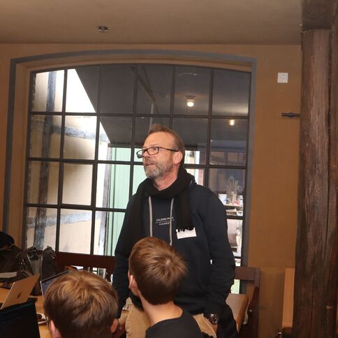 Ein Mann spricht im Tuppenhof Kaarst zu einer Gruppe in einem rustikalen Raum mit großen Fenstern.