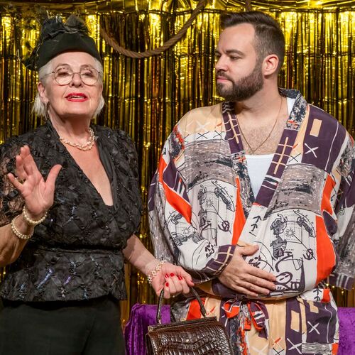 Eine ältere Frau und ein Mann in buntem Gewand stehen auf einer Theaterbühne vor einem goldenen Vorhang.