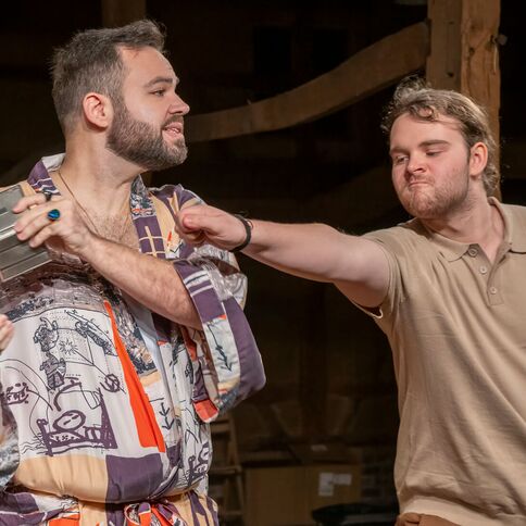 Zwei Männer auf einer Theaterbühne interagieren humorvoll, wobei einer einen schelmischen Gesichtsausdruck zeigt.