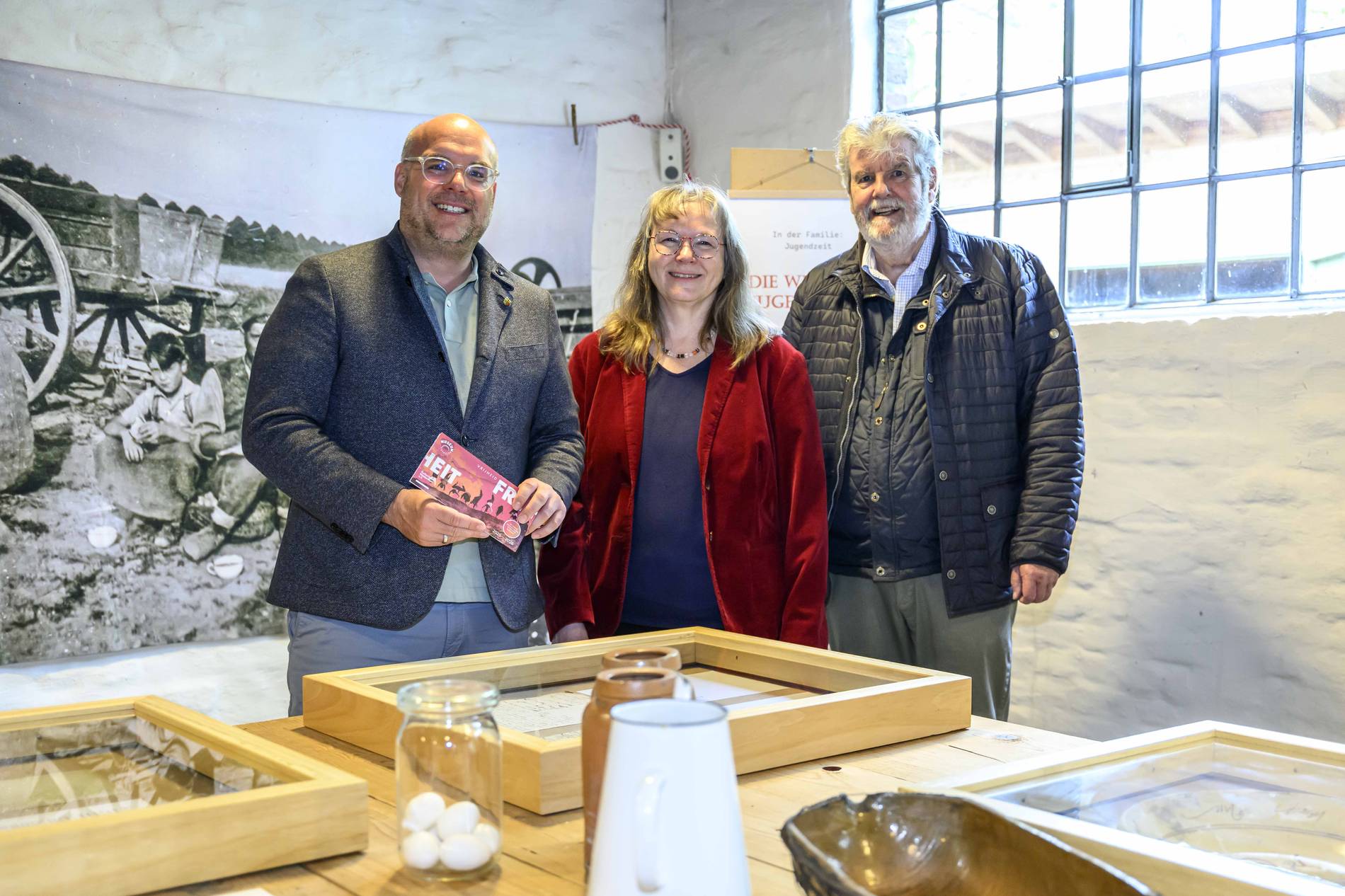 Drei Personen stehen im Tuppenhof, umgeben von Ausstellungsstücken und historischen Fotos aus Kaarst.